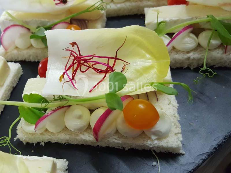 Tranche de pain garnie de fromage, légumes colorés, endive et germes, présentée sur une assiette noire.