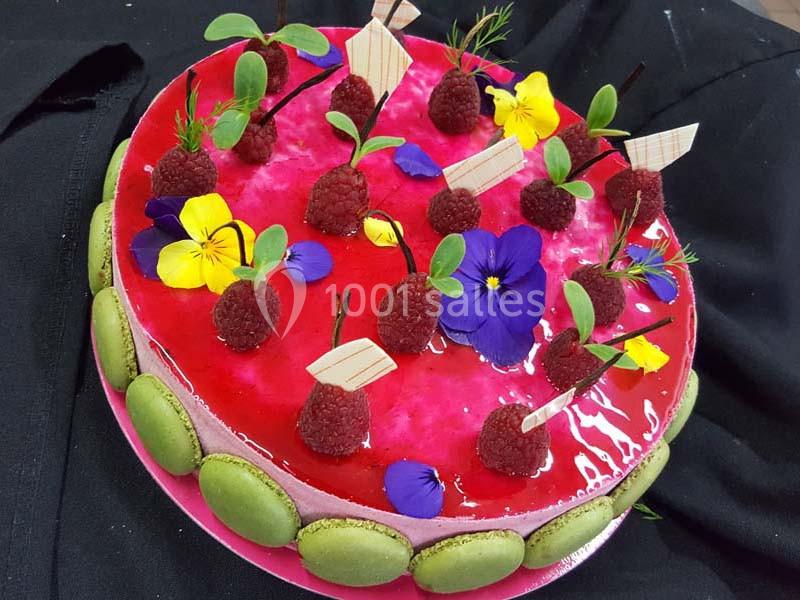 Gâteau décoré de framboises, fleurs comestibles, macarons verts et glaçage rouge brillant.