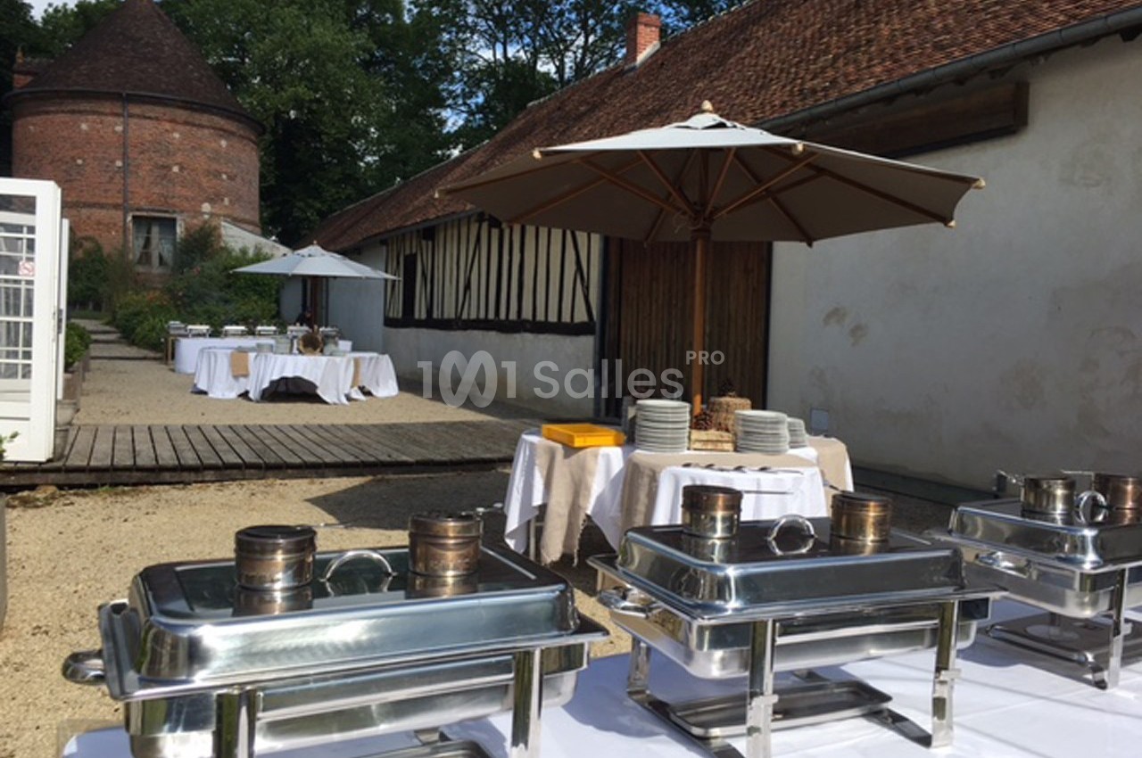 Buffet extérieur avec plats en inox, tables dressées et parasols dans une cour pavée près de bâtiments traditionnels.