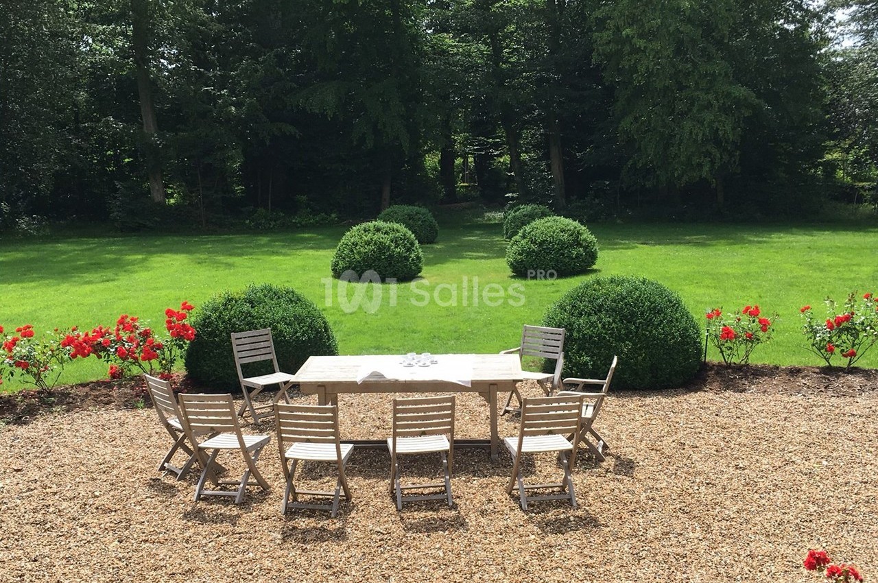 Table et chaises en bois disposées sur du gravier, face à un jardin verdoyant avec buissons et pelouse.