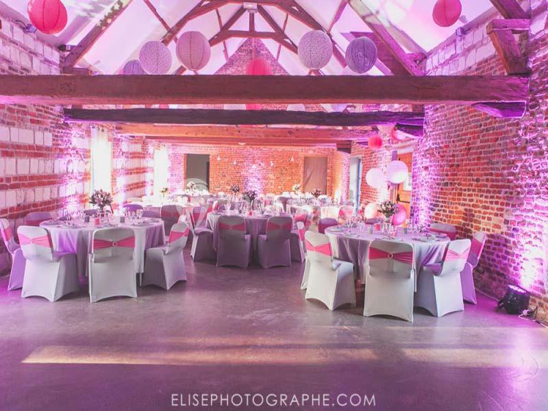 Salle de réception en briques avec tables rondes décorées, éclairage rose et lanternes suspendues.