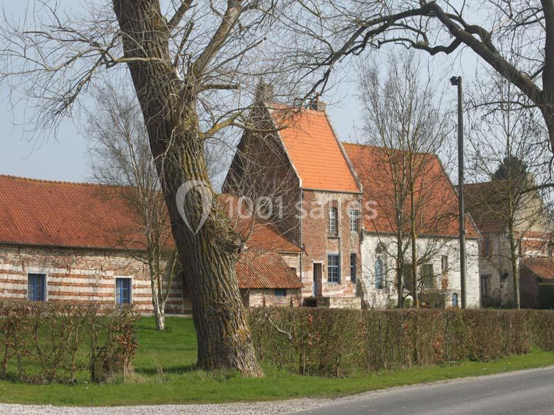 Ferme traditionnelle en briques et pierres avec toits en tuiles rouges, entourée d'arbres et d'une haie.