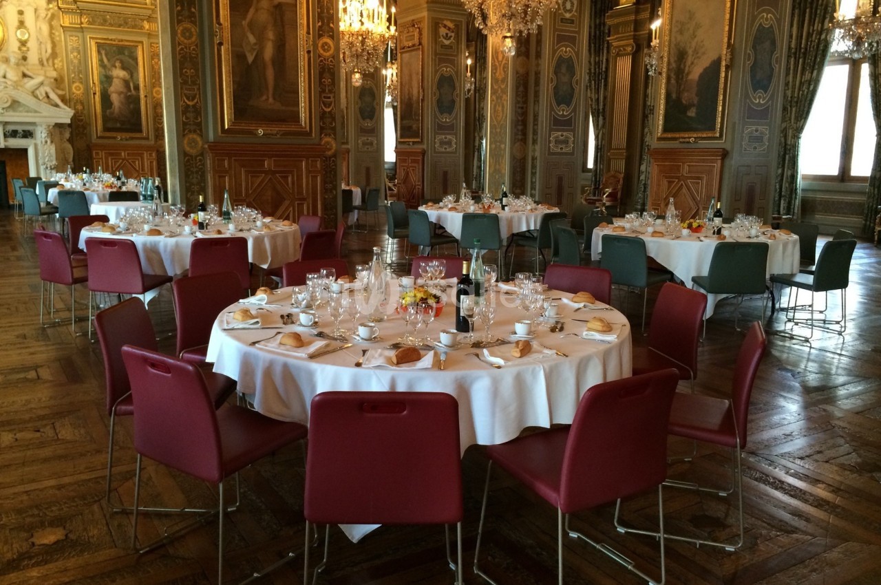 Salle élégante avec tables rondes dressées, chaises colorées, lustres et décorations murales ornées.