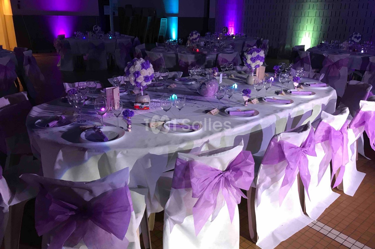 Salle de réception décorée avec des tables dressées et des chaises ornées de nœuds violets.