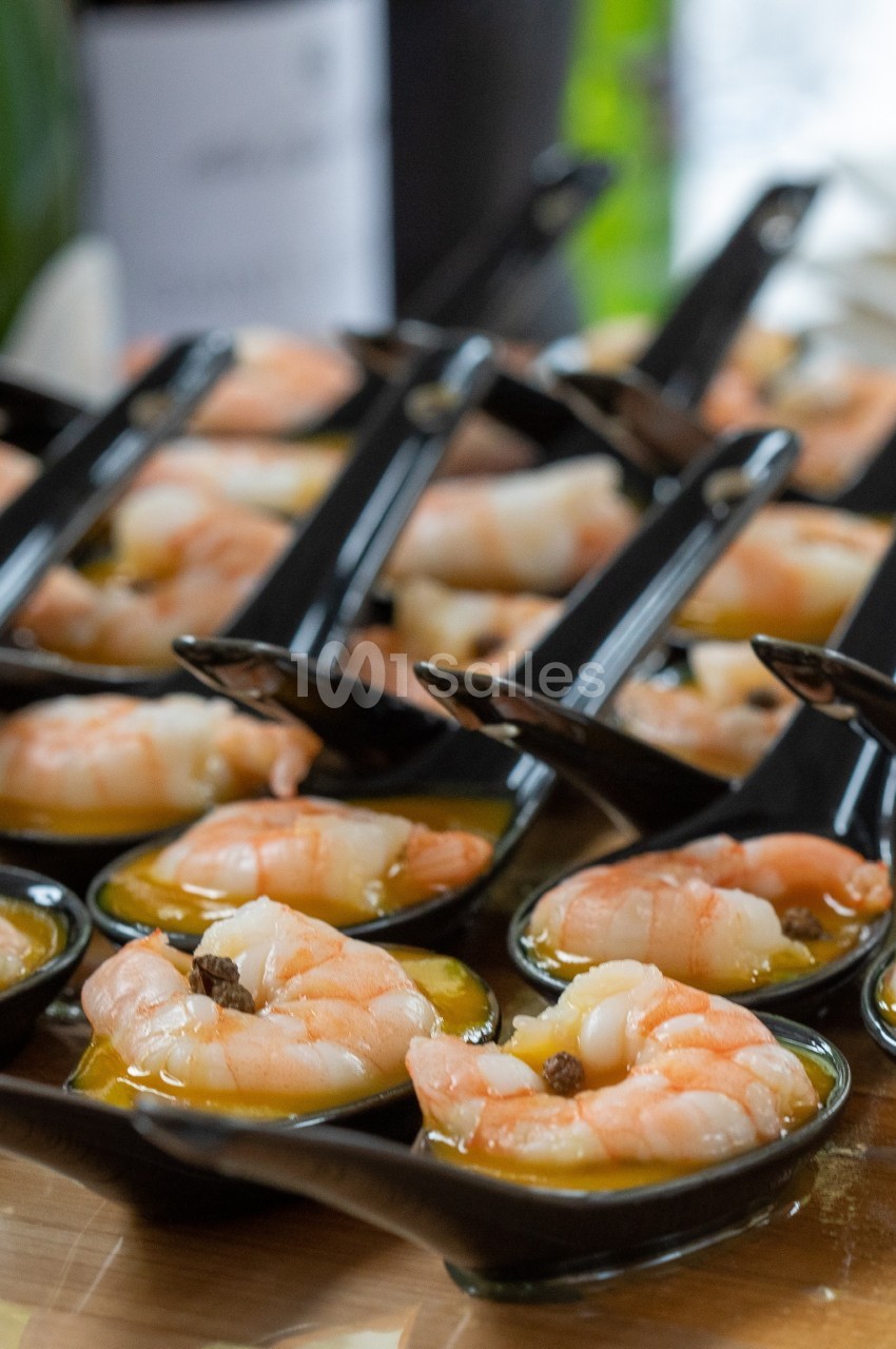 Crevettes servies dans des cuillères noires avec une sauce, présentées sur une table en bois.