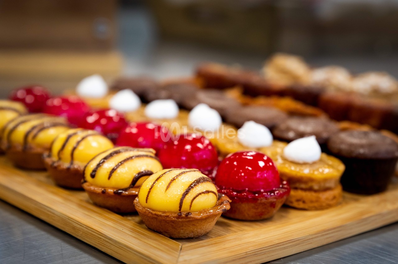 Plateau en bois garni de petites pâtisseries colorées, dont des tartelettes jaunes et rouges décorées de glaçage.