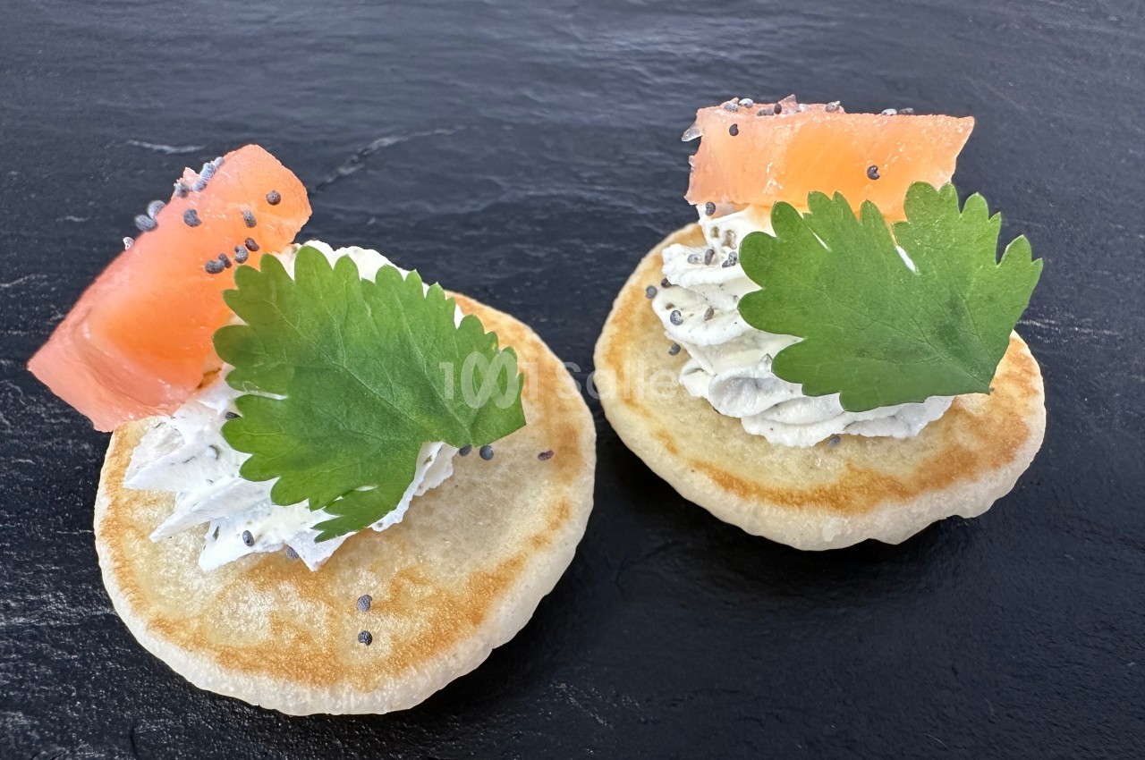 Deux blinis garnis de crème, saumon fumé, graines de pavot et feuilles de coriandre sur une ardoise noire.