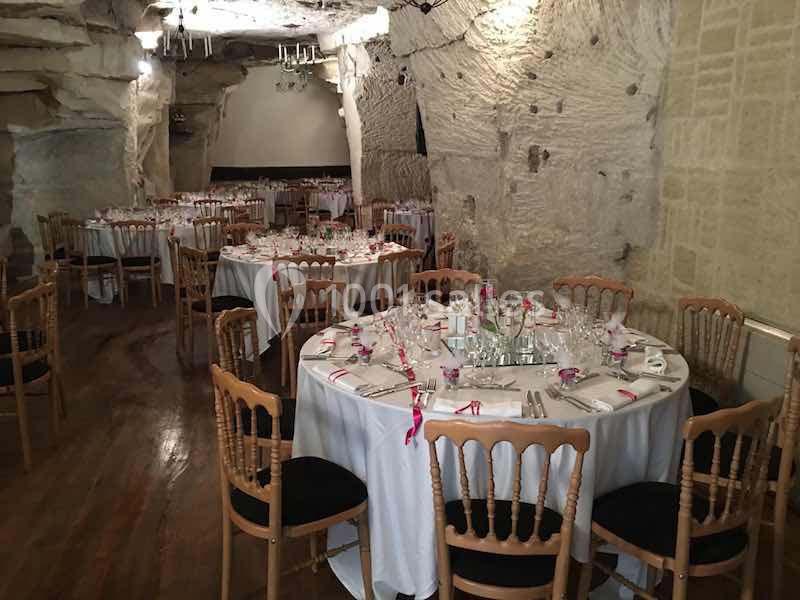 Salle de réception troglodytique avec tables rondes dressées, nappes blanches et chaises en bois.