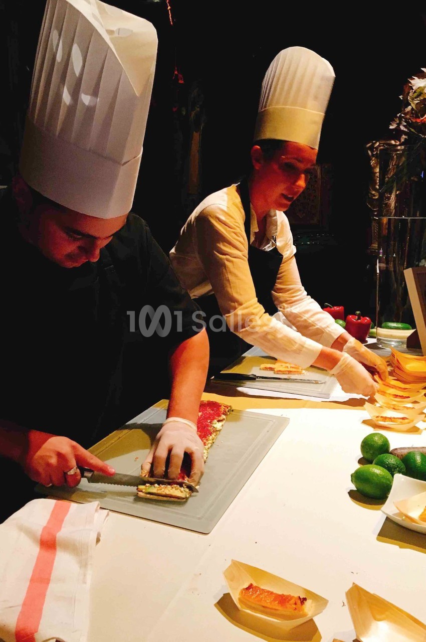Deux chefs en tenue blanche préparent des plats sur un plan de travail éclairé, entourés d'ingrédients frais.