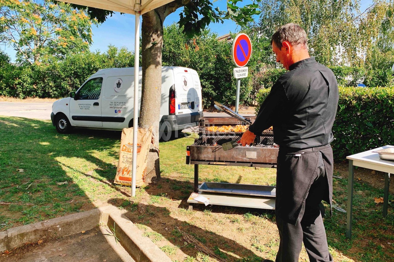 Un cuisinier en tenue noire prépare un barbecue en plein air près d'un véhicule utilitaire stationné.