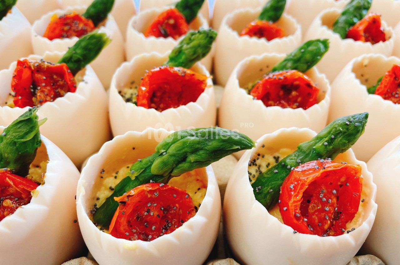 Œufs garnis de mousse, décorés de tomates cerises rôties et pointes d'asperges, présentés en rangées.