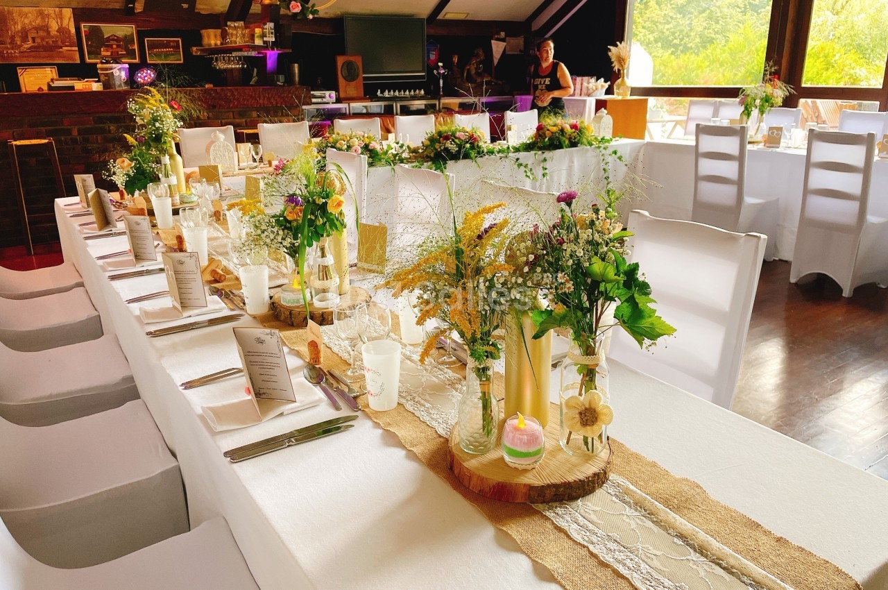 Table décorée pour un événement avec nappes blanches, fleurs, bougies et menus, dans une salle lumineuse.