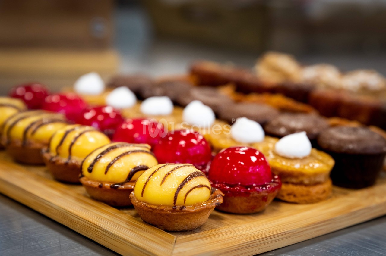 Plateau en bois garni de petites pâtisseries colorées, dont des tartelettes jaunes et rouges décorées de glaçage.