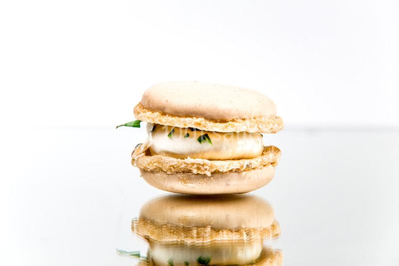 Macaron beige garni de crème blanche et décoré de petites herbes, posé sur une surface réfléchissante.