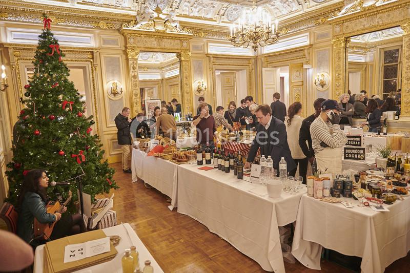 Salle élégante avec des stands de produits artisanaux, un sapin décoré et des visiteurs échangeant autour des tables.