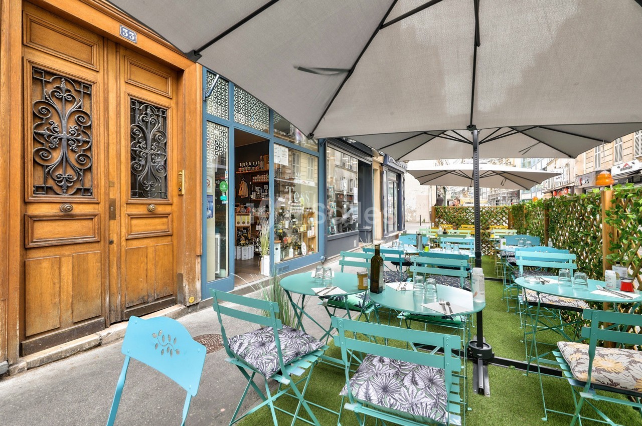 Terrasse extérieure avec tables et chaises turquoise, parasols, et entrée d'un commerce en arrière-plan.