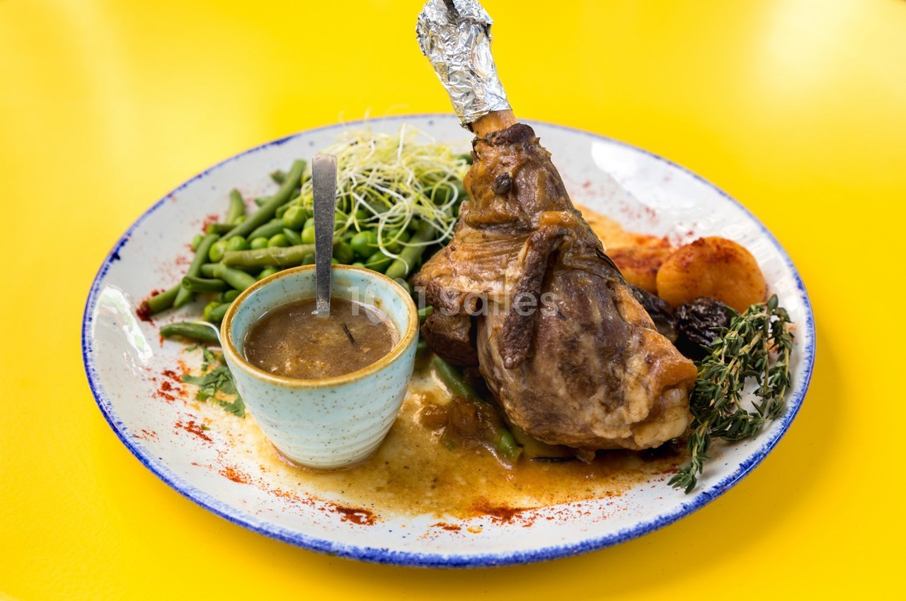 Assiette avec un jarret d'agneau, haricots verts, purée, pruneaux, sauce en bol et herbes, sur fond jaune.