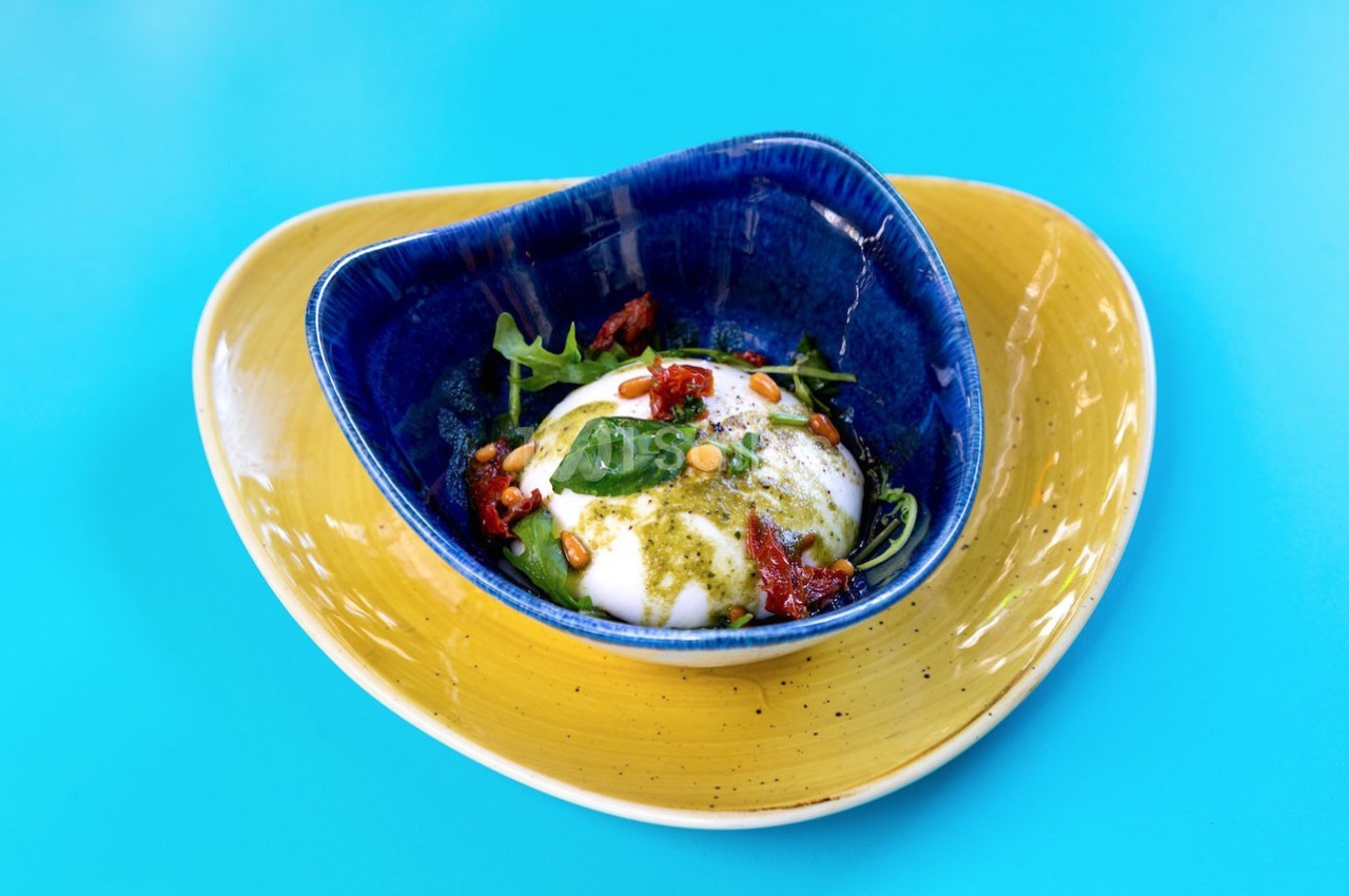 Assiette colorée contenant une burrata garnie de tomates séchées, pignons, roquette et pesto, sur fond bleu vif.