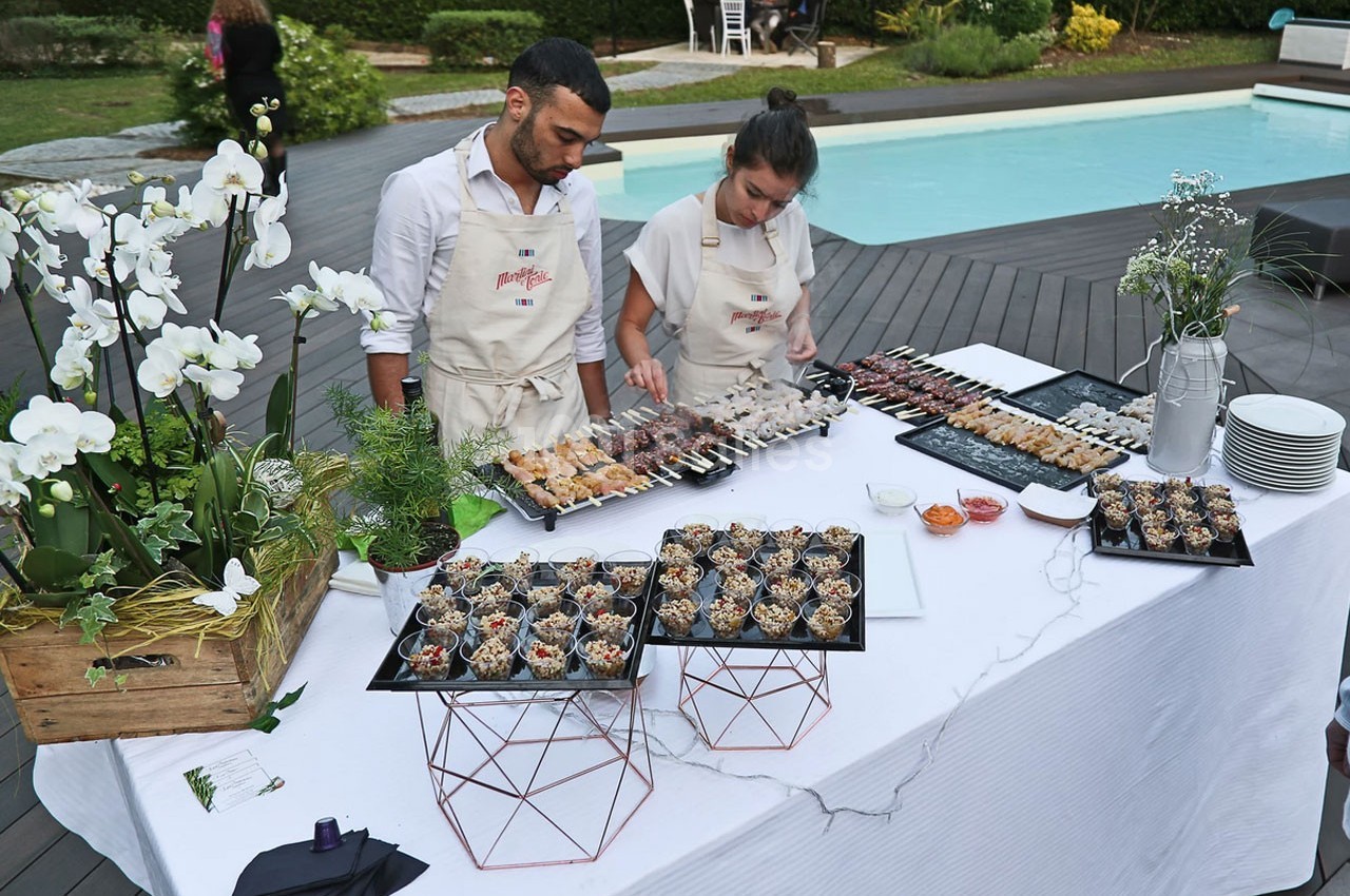 Deux personnes préparent un buffet de brochettes et verrines sur une table dressée près d'une piscine.