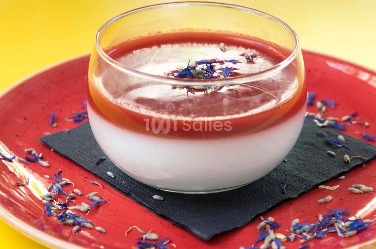 Dessert en verrine avec une couche blanche, un coulis rouge et des fleurs comestibles sur une assiette rouge.