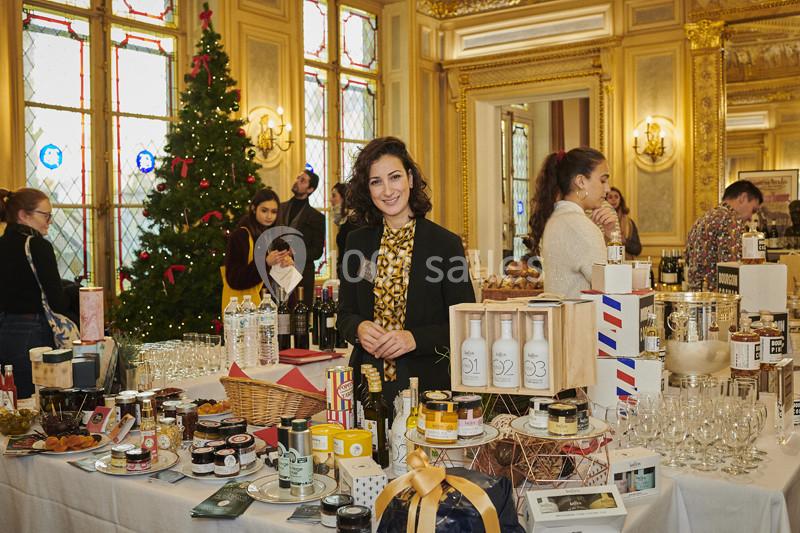 Une femme souriante se tient derrière un stand de produits variés dans une salle décorée avec un sapin de Noël.