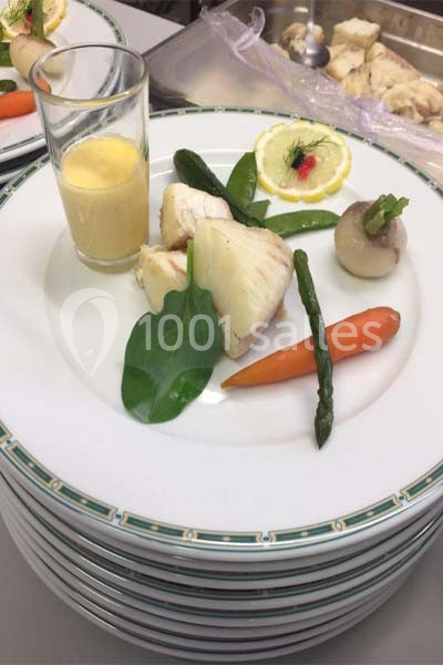 Assiette dressée avec poisson, légumes vapeur, sauce en verrine et tranche de citron décorée, sur une pile d'assiettes.