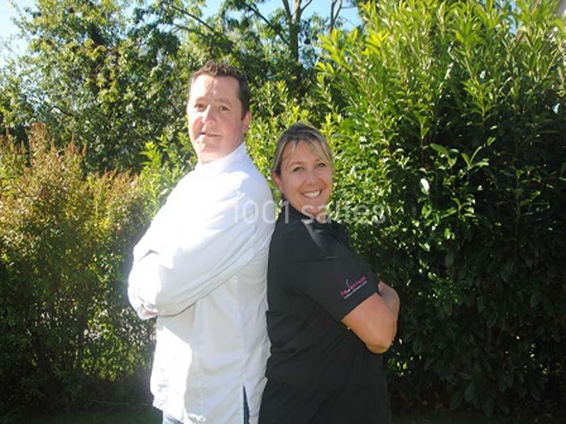 Deux personnes en tenue professionnelle, dos à dos, souriant dans un jardin ensoleillé avec des arbres en arrière-plan.