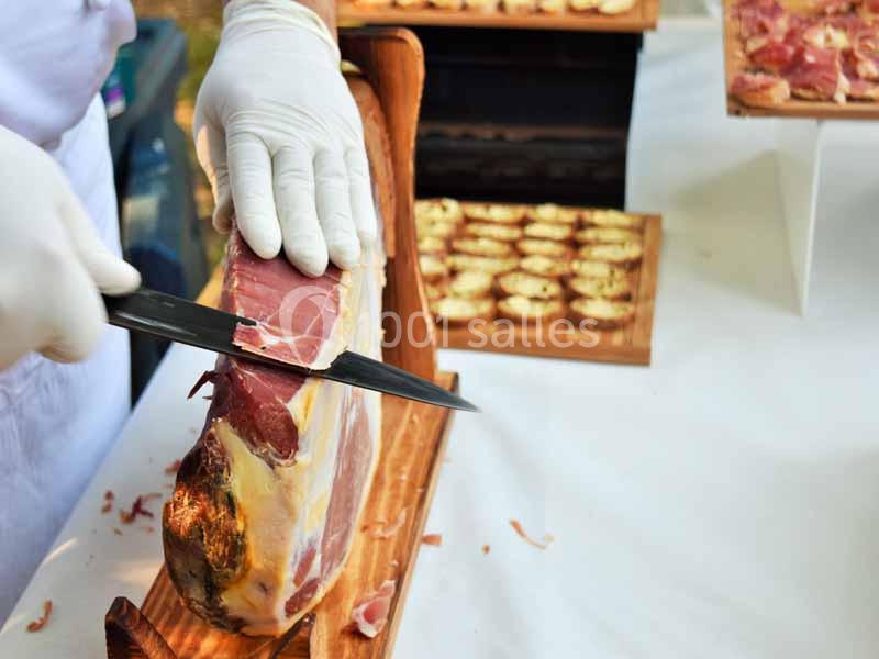 Un cuisinier découpe finement du jambon cru sur un support en bois, avec des amuse-bouches en arrière-plan.