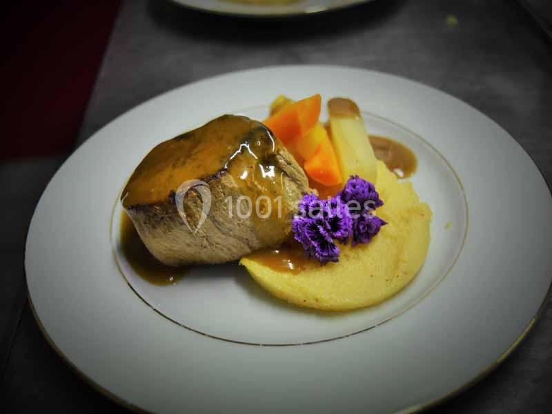 Assiette contenant un filet de viande nappé de sauce, accompagné de légumes, purée et fleurs comestibles violettes.