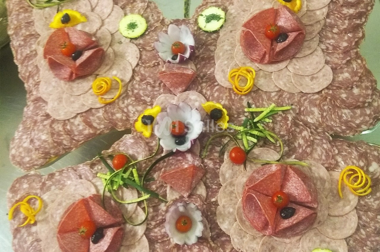 Plateau de charcuterie décoratif avec des tranches de saucisson, fleurs en légumes et garnitures colorées.