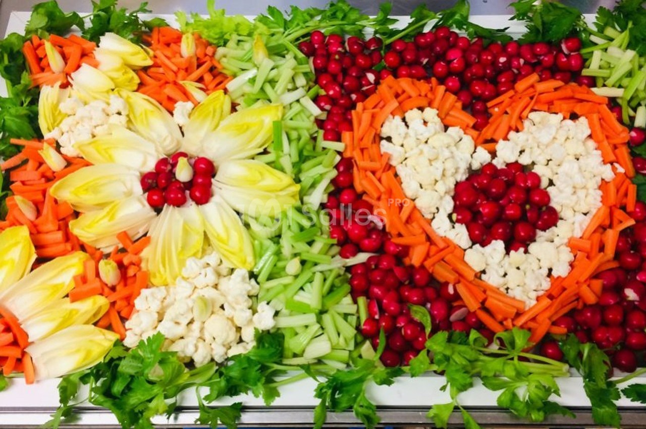 Plateau de crudités coloré disposé en motifs floraux et en cœur, avec carottes, céleri, endives, chou-fleur et canneberges.