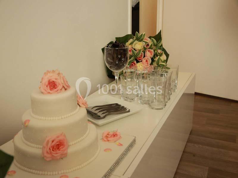 Gâteau blanc décoré de roses, verres et couverts disposés sur un meuble blanc dans une pièce au sol en bois.