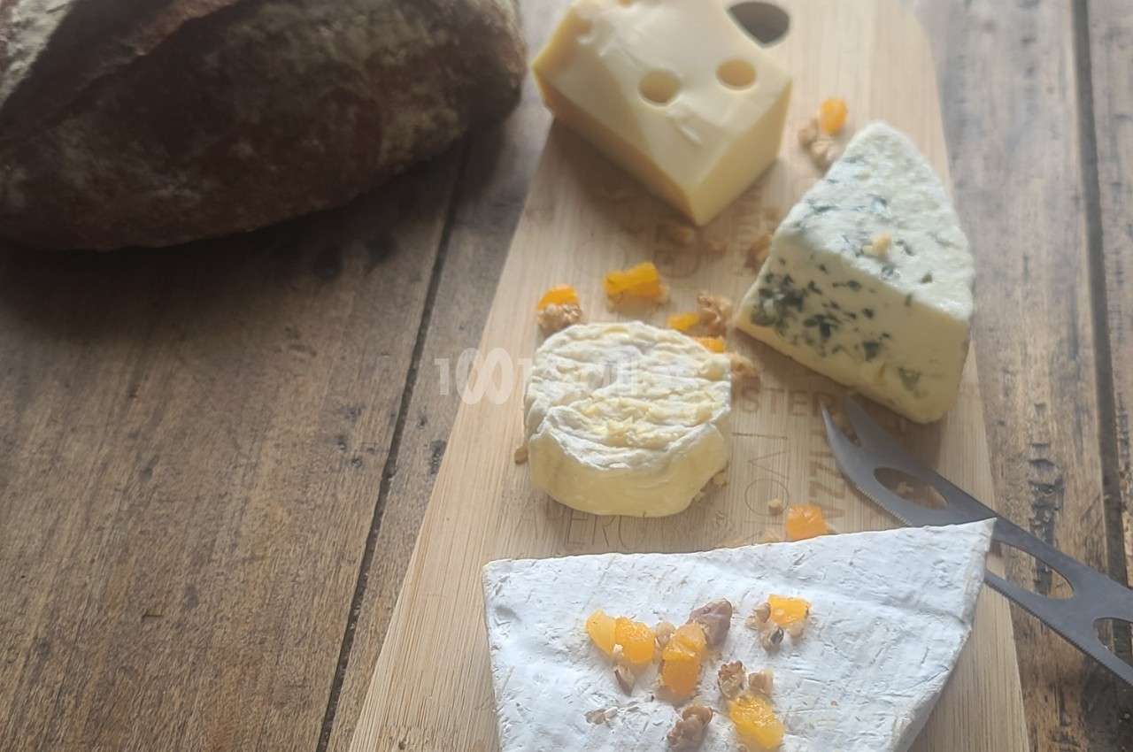 Plateau en bois avec quatre fromages variés, morceaux d'abricots secs et pain en arrière-plan sur une table en bois.