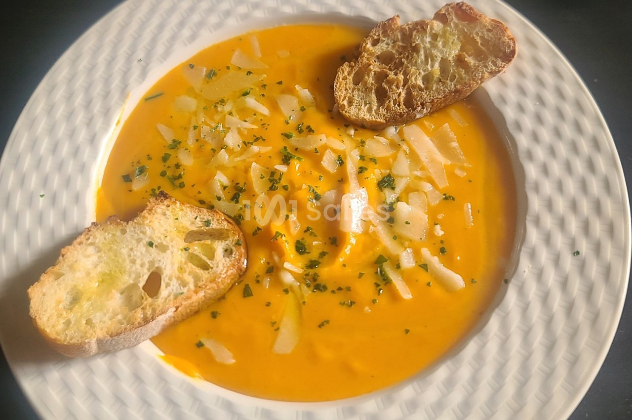 Soupe orange garnie de copeaux de fromage, herbes et accompagnée de deux tranches de pain grillé.