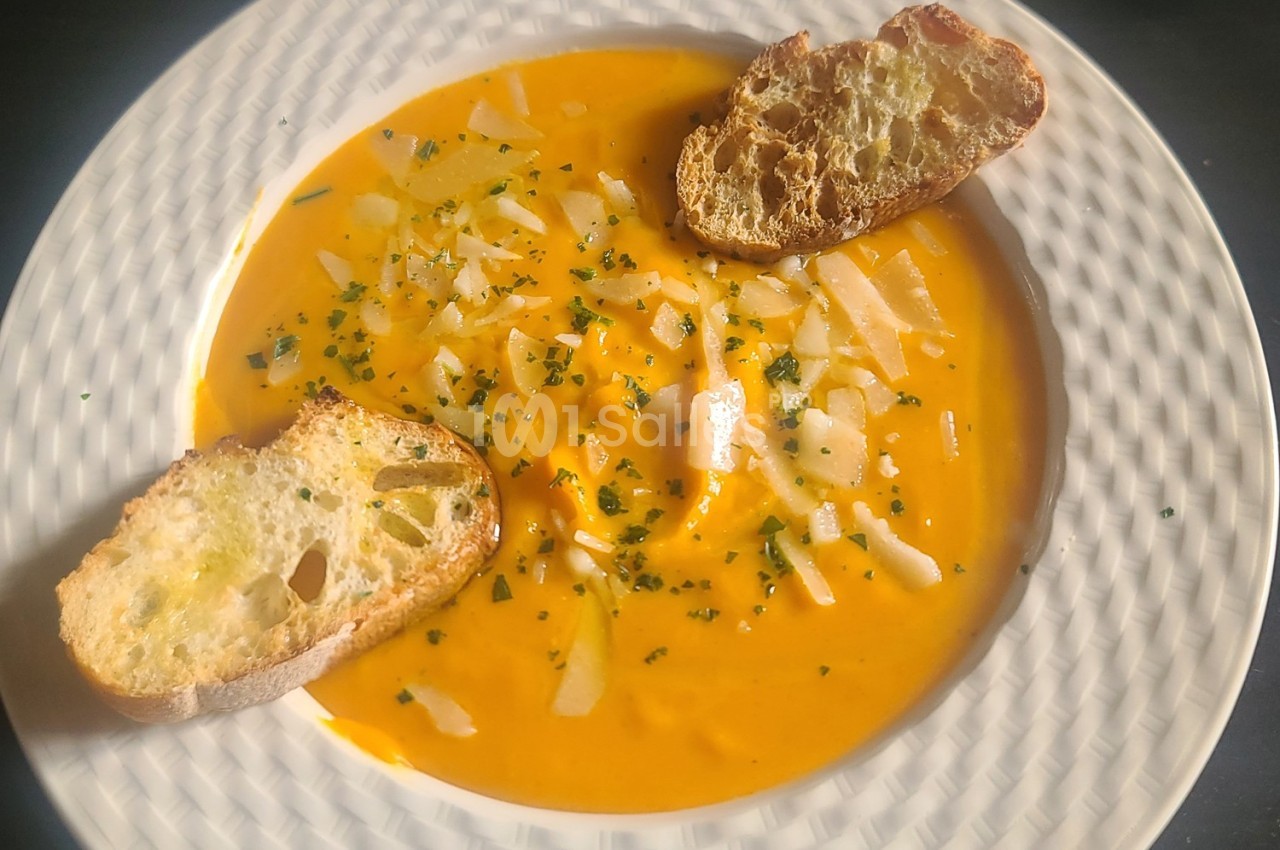 Soupe orange garnie de copeaux de fromage, herbes et accompagnée de deux tranches de pain grillé.