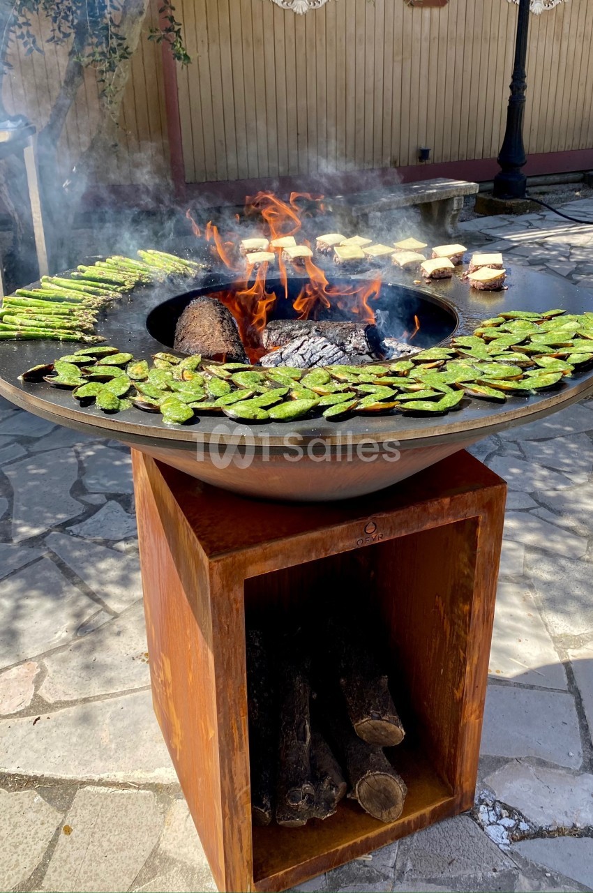 Grill circulaire avec des légumes verts et des aliments en cuisson sur un feu de bois dans un espace extérieur ensoleillé.
