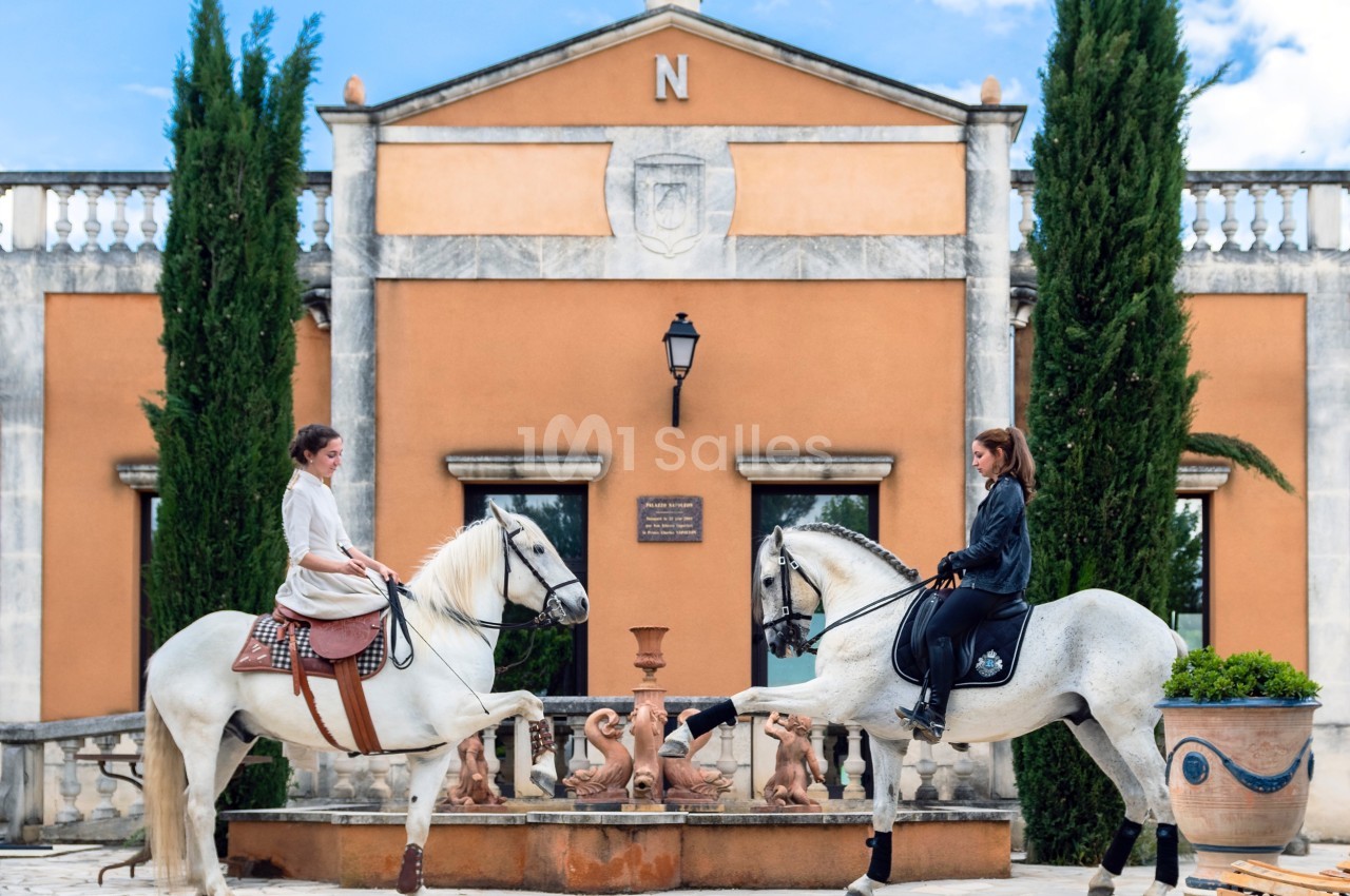 Deux cavalières à cheval se saluent devant une fontaine et un bâtiment à l'architecture classique.