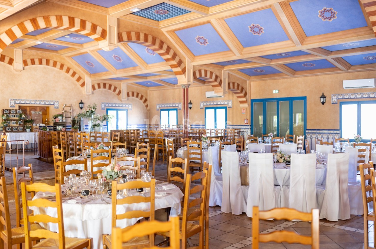 Salle de réception lumineuse avec tables rondes dressées, chaises en bois et plafond décoré de motifs colorés.