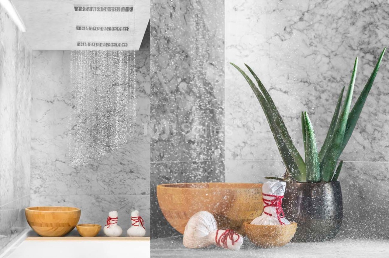Douche moderne avec mur en marbre, bols en bois, plantes et accessoires de spa disposés sur une surface.