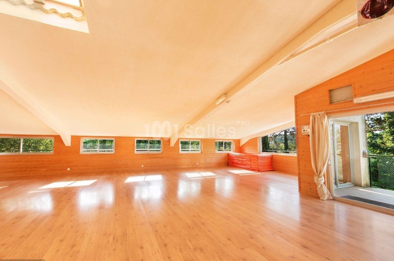 Salle spacieuse avec parquet clair, murs en bois, grandes fenêtres et lumière naturelle, donnant sur un espace extérieur.