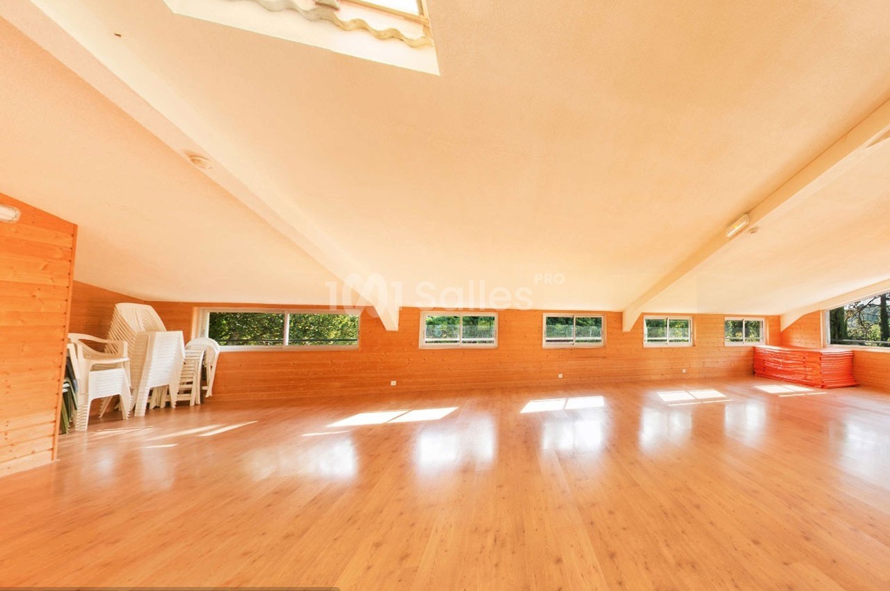 Salle lumineuse avec parquet en bois, murs en bois clair, grandes fenêtres et chaises empilées dans un coin.