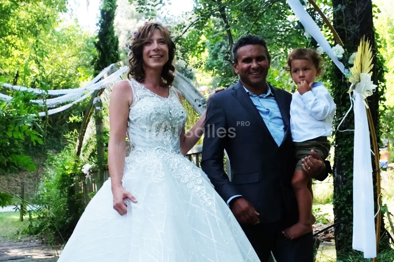 Un couple en tenue de mariage pose avec un enfant dans un jardin décoré de rubans et de fleurs.