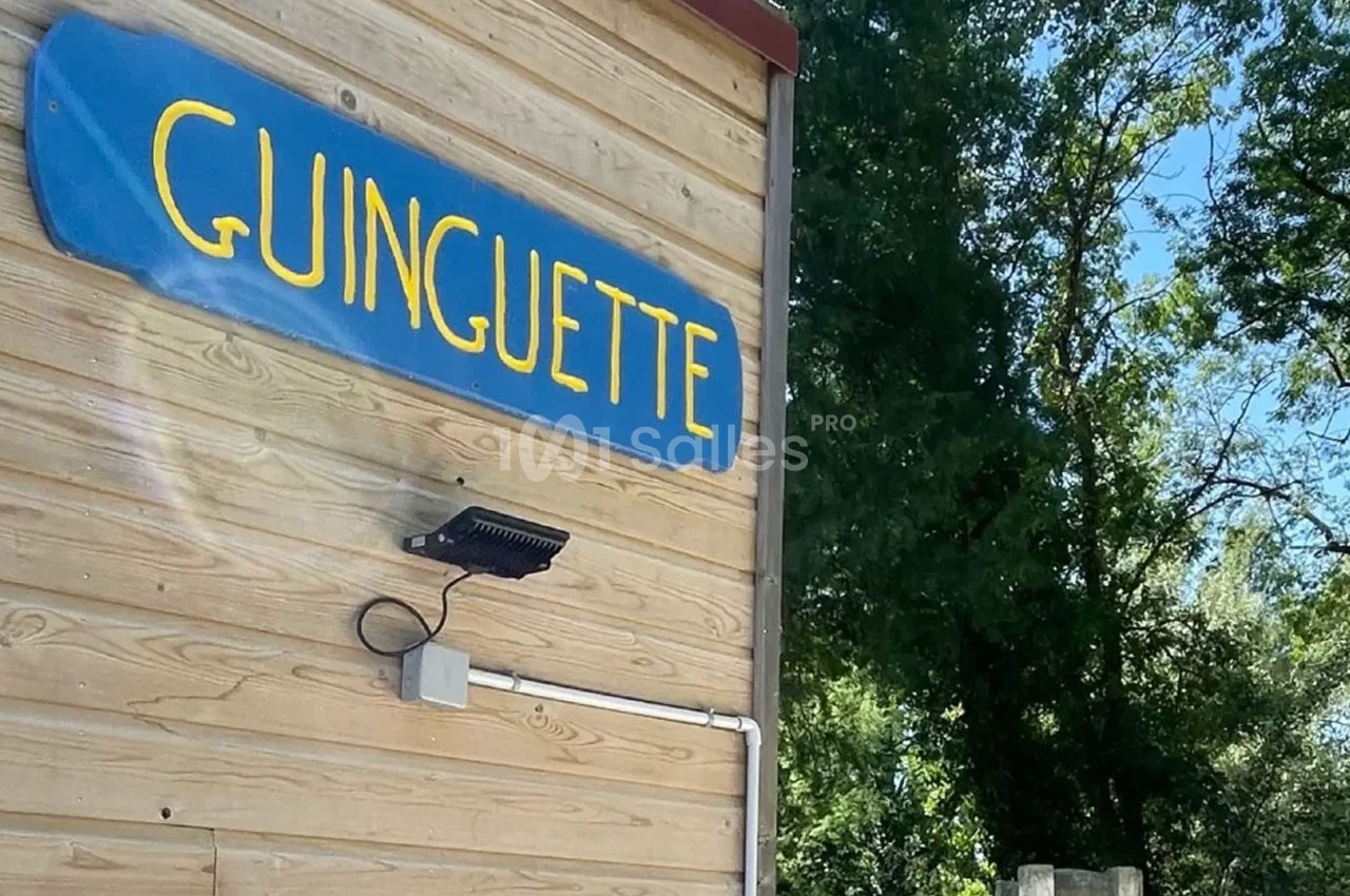 Façade en bois avec une enseigne bleue indiquant ’Guinguette’, entourée de végétation en arrière-plan.