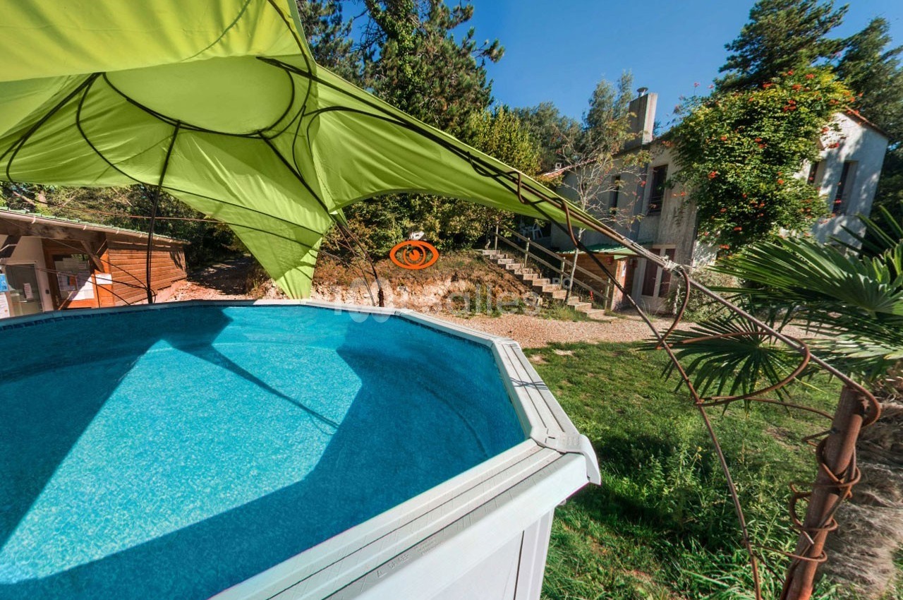 Piscine hors-sol entourée de verdure avec une maison en arrière-plan et une voile d'ombrage verte au-dessus.