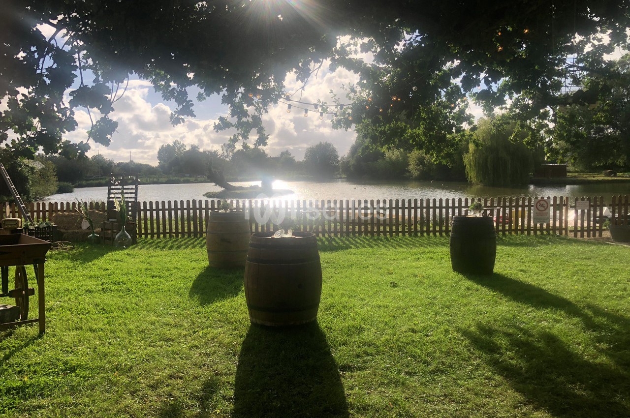 Vue d'un jardin ombragé avec des tonneaux en bois, une clôture en bois et un étang en arrière-plan sous un ciel ensoleillé.