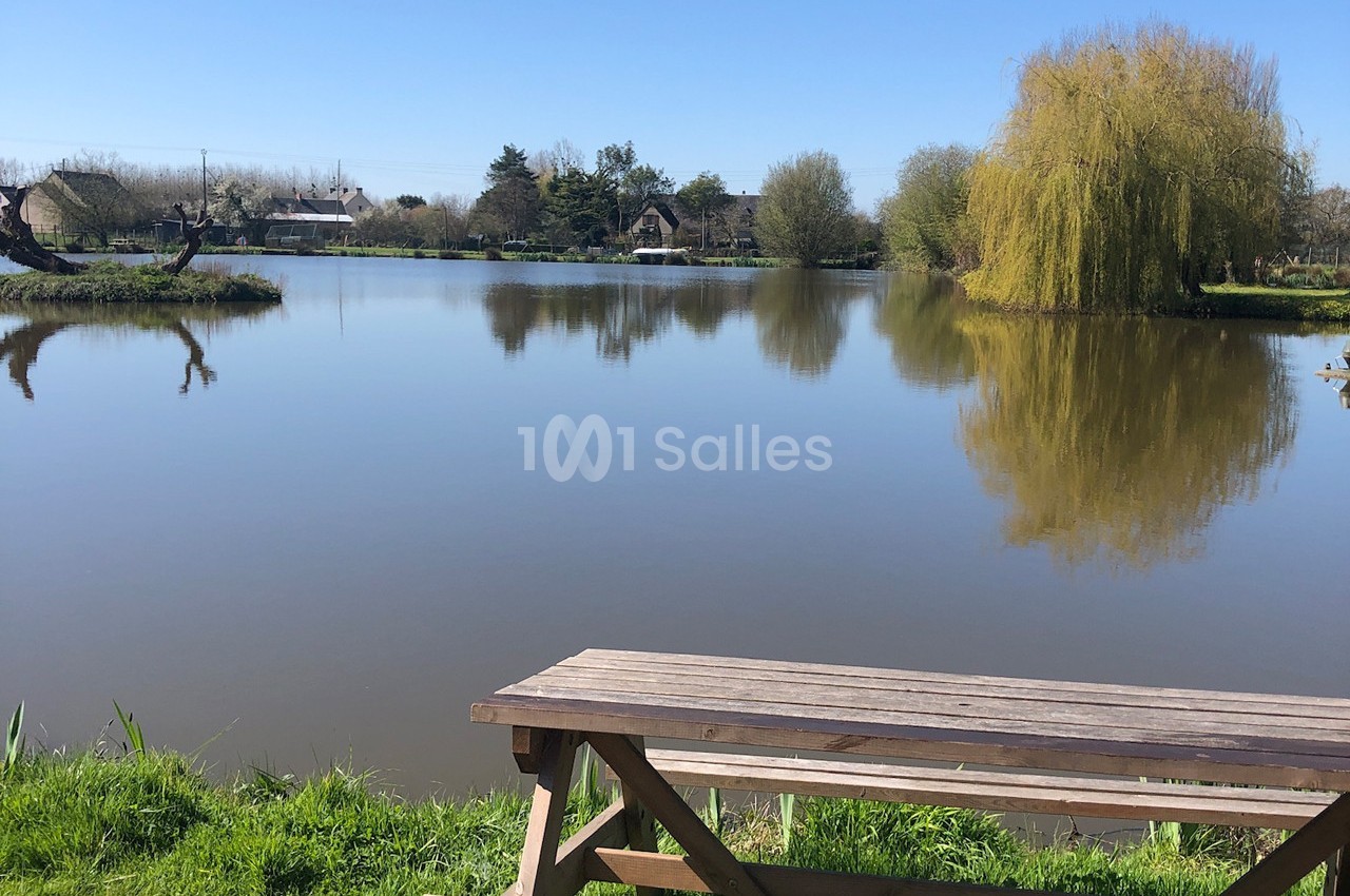 Vue d'un étang calme entouré d'arbres sous un ciel bleu, avec une table de pique-nique en bois au premier plan.