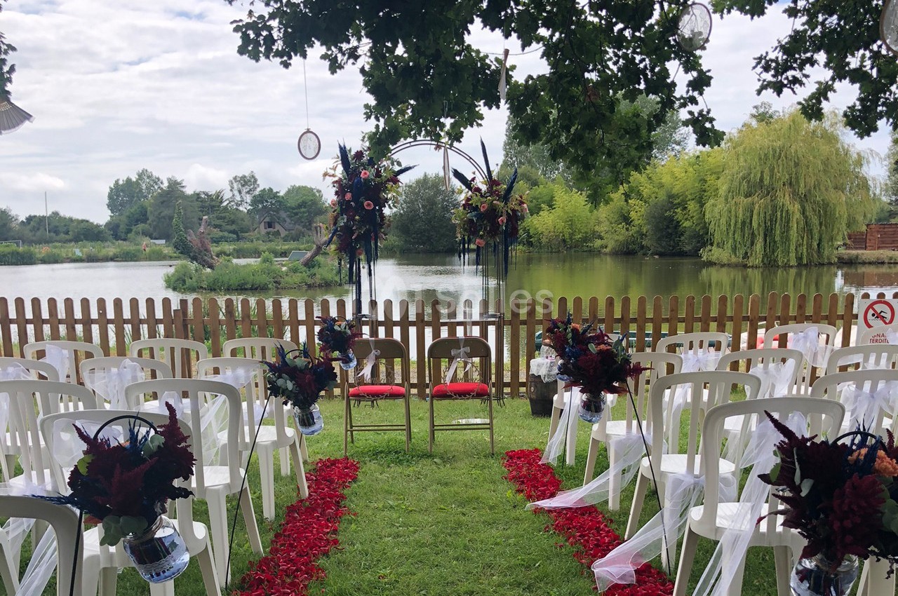 Chaises blanches alignées devant une arche fleurie, disposées pour une cérémonie en plein air près d'un étang.