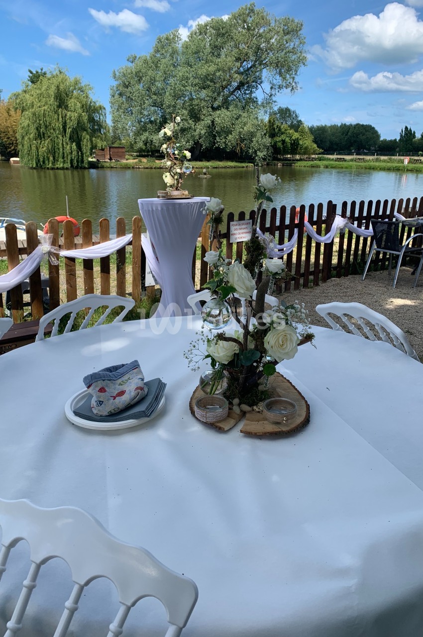Table décorée avec des fleurs blanches et des rondins de bois, près d'un lac entouré de verdure et d'une clôture en bois.