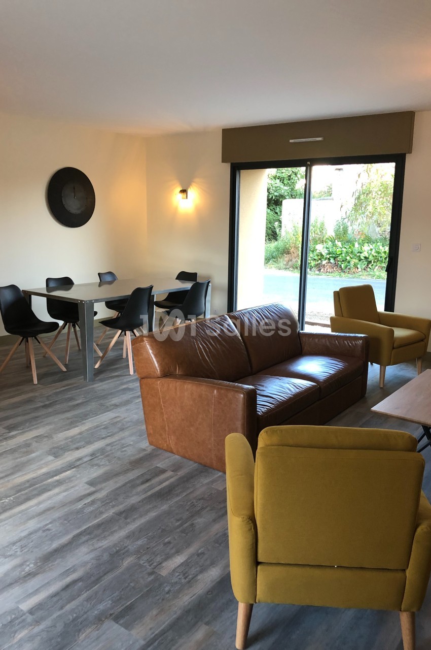 Salon lumineux avec canapé en cuir, fauteuils jaunes, table à manger et vue sur un jardin par une baie vitrée.