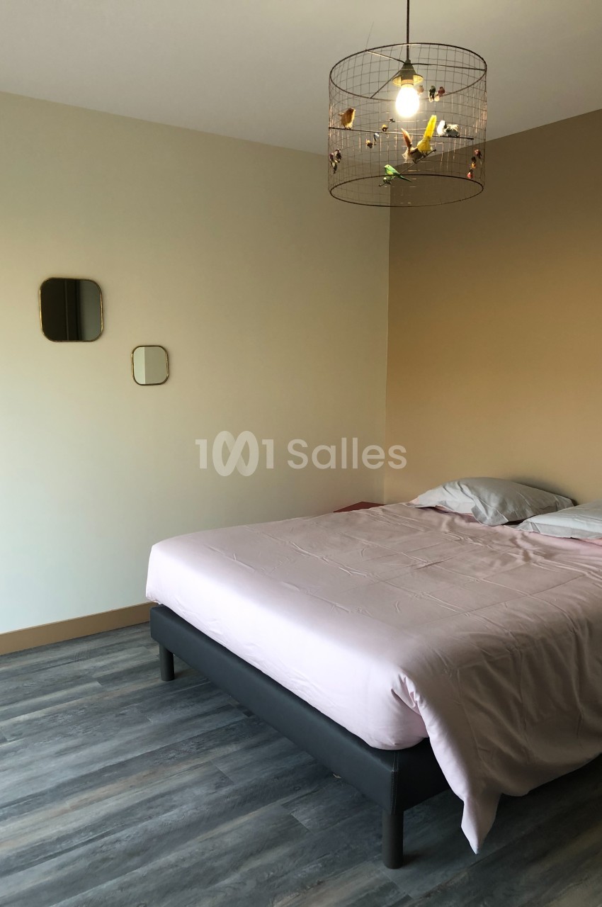 Chambre avec un lit double aux draps roses, sol en parquet gris, murs beige clair et suspension en métal décorative.